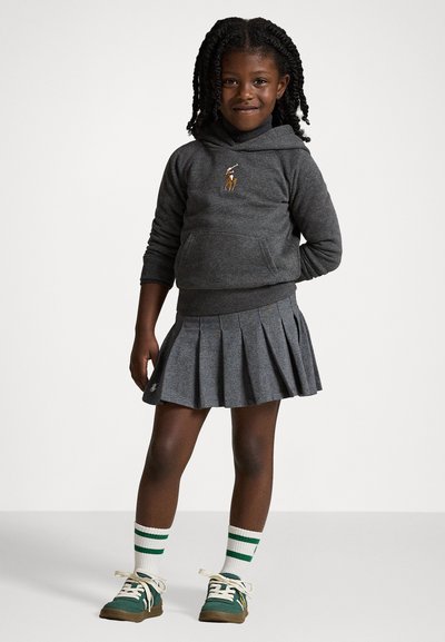 Γκρίζα φούτερ με λογότυπο, πλισέ γκρι φούστα, πράσινα αθλητικά παπούτσια με λευκά κορδόνια και κάλτσες μέχρι το γόνατο με πράσινες ρίγες, σε ελαφρύ φόντο.