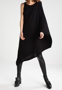 Svart asymmetrisk klänning i mjukt tyg, med enaxlad design och flödande silhuett, kombinerad med tajta svarta leggings och ankelstövlar.