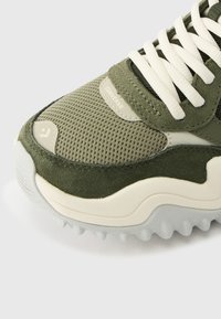 Κοντινό πλάνο πράσινου ελιάς αθλητικού παπουτσιού Converse με ύφασμα δίχτυ, πάνω μέρη από σουέντ, λευκά κορδόνια και υφή λευκής σόλας με λογότυπο αστέρι στη μύτη.