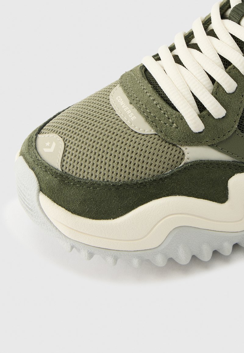 Nahaufnahme eines olivgrünen Converse-Sneakers mit Mesh-Gewebe, Wildleder-Overlays, weißen Schnürsenkeln und strukturierter weißer Sohle mit Sternlogo an der Spitze.