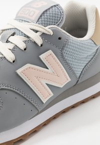 Zapatilla gris con parte superior de malla y cuero, con un logo "N" rosa destacado, cordones blancos y suela de goma. Se aprecian detalles texturizados.