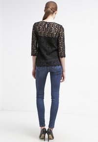 Top in pizzo nero con maniche a tre quarti, caratterizzato da un dettaglio a chiave sulla schiena, abbinato a jeans in denim scuro e tacchi neri.