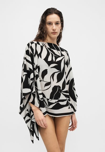 Νεαρή γυναίκα με βρεγμένα μαλλιά, που φοράει ένα μαύρο και άσπρο χαλαρό τοπ με έντονα αφηρημένα φυλλώδη σχέδια πάνω σε απλό ανοιχτό φόντο.