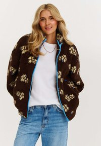 Hnědá fleecová bunda s květinovými vzory, světle modrý zip, stojáčkový límec a volný střih, navrstvená přes bílou košili a modré džíny.