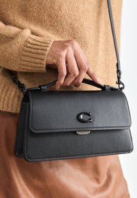 Bolso de mano de piel negra con cierre de solapa, textura suave, detalle del logotipo en relieve y una correa desmontable, sostenido contra una falda marrón.