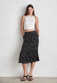 Falda negra con flores y dobladillo acampanado, que presenta flores blancas y amarillas. Combinada con una blusa ajustada blanca sin mangas y sandalias de tacón negras.