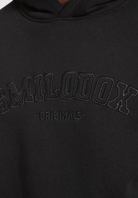 Felpa nera con cappuccio, con ricamo in rilievo nero che recita "SMILODOX ORIGINALS." È realizzata in tessuto morbido e ha una vestibilità standard.