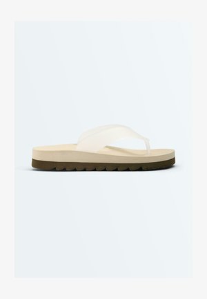 Beige teenslipper met dikke zool, witte Y-vormige band en donkerbruine textuurzool, zijaanzicht op een witte achtergrond.