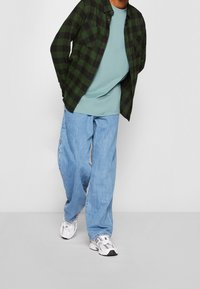 Grön och svart rutigt button-up skjorta över en ljusblå t-shirt, parat med lösa ljusblå jeans och vita sneakers.