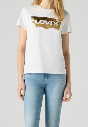 Biała bawełniana koszulka z krótkim rękawem i okrągłym dekoltem, z złotym foliowym logo "Levi's" na piersi, w zestawieniu z jasnoniebieskimi dżinsami.