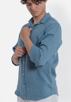 Giovane uomo con capelli ricci che allaccia una camicia blu a maniche lunghe, guardando verso il basso, su sfondo bianco uniforme.