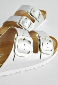 Silverfärgade sandaler med två breda remmar, utrustade med stora fyrkantiga metallspännen. Mjuk beige fotbädd och vit gummiyttersula.