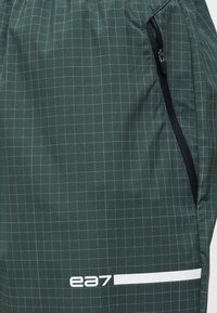 Σκούρο πράσινο ύφασμα με ανοιχτόχρωμο μοτίβο πλέγματος, μαύρο φερμουάρ στη δεξιά πλευρά και λευκό λογότυπο "ea7" με οριζόντιο μπάρ κοντά στο κάτω μέρος.