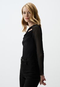 Schwarzes Langarmshirt mit einem drapierten Ausschnitt und durchsichtigen Ärmeln, kombiniert mit hochtaillierten schwarzen Jeans mit dezenten Taschendetails.