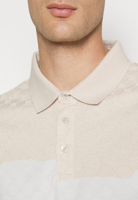Camisa polo beige con un patrón texturizado en la sección superior, que presenta un cuello clásico y cinco botones, y una tela suave en la parte inferior.