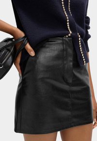 Osoba v černé kožené mini sukni a námořnickém modrém svetru s kamínkovým lemem, držící malou černou koženou tašku.