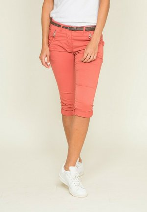 Personne portant un pantalon corail roulé aux genoux avec une ceinture marron et des baskets blanches, se tenant devant un fond clair uni.