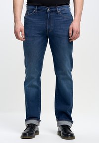 Vaqueros de mezclilla azul oscuro con corte recto, costuras visibles, cinco bolsillos y puños enrollados, combinados con zapatos de cuero negros.
