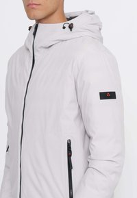 Giacca grigia chiaro con cerniera e cappuccio, con zip nere, tasca laterale e etichetta con logo sulla manica. Texture liscia.