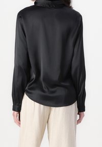 Donna che indossa una blusa in satin nera a maniche lunghe e pantaloni beige larghi, vista di spalle su uno sfondo bianco uniforme.