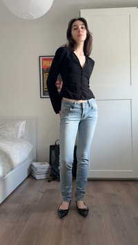 Nainen mustassa napitetussa paidassa ja vaaleansinisissä farkuissa seisoo makuuhuoneessa, jossa hänen takanaan on sänky, kehystetty juliste ja valkoinen vaatekaappi.