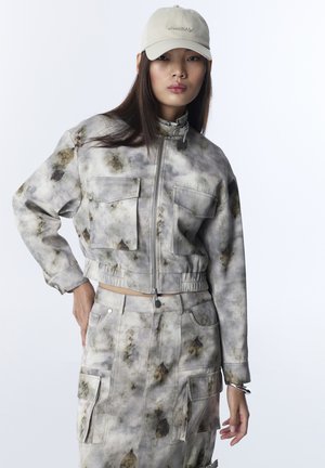 Kobieta w beżowym zestawie z kurtką i spódnicą w stylu tie-dye z dużymi kieszeniami oraz beżową czapką, pozująca na tle białego, jednolitego tła.