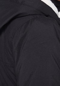 Svart jacka med en texturerad, matt tyg. Har en foderhuva och synliga sömdetaljer. Nära bild av axelområdet.