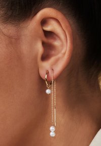 Boucle d'oreille en or avec une chaîne, deux perles blanches et un pendentif en barre droite. Gros plan sur le détail de l'oreille.