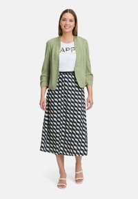 Veste vert clair sur un t-shirt blanc avec un motif graphique, associée à une jupe maxi à motifs ondulés bleu marine et crème et des talons beiges.