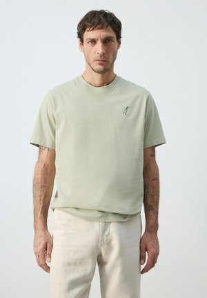 Bărbat cu păr scurt și închis la culoare, purtând un tricou verde pal și pantaloni de culoare alb-crem, stând pe un fundal simplu, deschis.