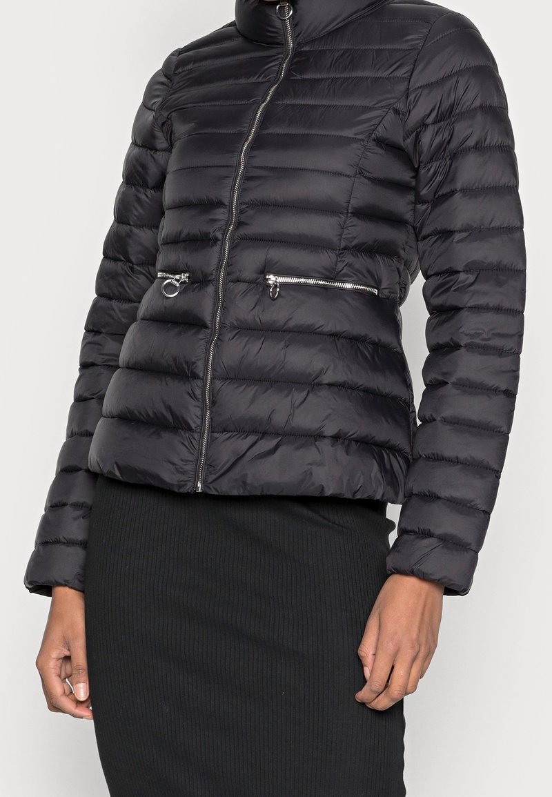 Svart puffjacka med hög krage, horisontellt quiltat mönster och två framfickor med dragkedja. Dragkedjestängning med metalliska detaljer.