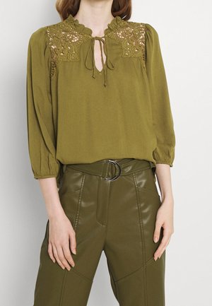 Mujer que lleva una blusa verde oliva con detalle de encaje en los hombros y el escote, combinada con pantalones verde oliva de talle alto con cinturón con anilla plateada.