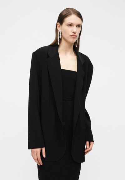 Γυναίκα με ίσια καστανά μαλλιά φοράει χοντρό μαύρο σακάκι πάνω από αμάνικο μαύρο φόρεμα, διακοσμημένο με μακριά, κρεμαστά σκουλαρίκια με στρας.