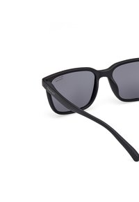 Gafas de sol negras con un marco geométrico y lentes grises, presentan un acabado mate y un brazo lateral de marca con la inscripción "GANT".