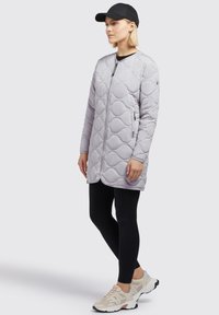 Veste longue matelassée gris clair avec des formes arrondies, fermeture éclair à l'avant, poches latérales et coupe décontractée, associée à des leggings noirs et des baskets beiges.