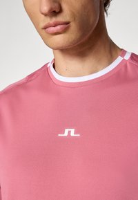 Rózsaszín sportpóló kerek fehér gallérral és diszkrét kontrasztcsíkokkal. A mellkason egy kis fehér logo található. Puha tapintású.
