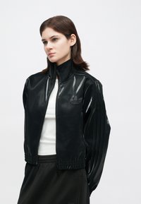 Γυναίκα με καστανά μαλλιά που φτάνουν μέχρι τους ώμους, φορώντας μια γυαλιστερή μαύρη τζάκετ με φερμουάρ πάνω από ένα λευκό μπλουζάκι και μαύρο παντελόνι, σε έναν απλό φωτεινό φόντο.