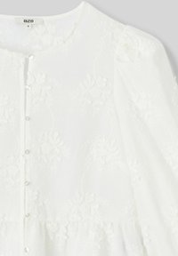 Blouse blanche avec broderie florale, fermeture par boutons, col rond et manches bouffantes. Fabriquée en tissu léger avec une texture douce.