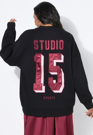 Persoon draagt een zwarte oversized trui met "STUDIO 15 SPORTS" in grote kastanjebruine letters en cijfers op de achterkant.