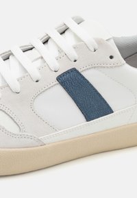 Zapatillas blancas con detalles de ante gris y una franja texturizada azul. Presentan cordones blancos y una suela de goma de color marrón claro con un acabado texturizado.