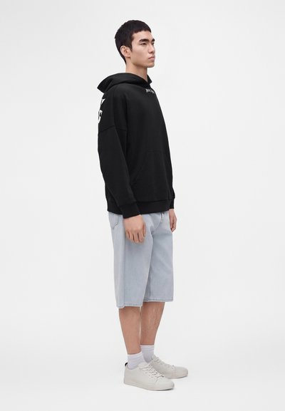 Jeune homme debout de profil, portant un sweat à capuche noir, un short en jean bleu clair jusqu'aux genoux, des chaussettes blanches et des baskets blanches.