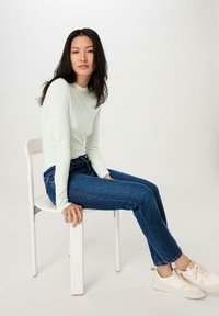 Hellgrünes Langarmoberteil, schlanker Schnitt; dunkelblaue Jeans; sitzt auf einem weißen Stuhl; cremefarbene Sneakers; glattes Material und sauberes Design.