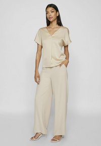 Bežinė satino palaidinė su V formos iškirpte ir trumpomis rankovėmis, derinama su plačiais bežiniais kelnais, pasižyminčiais lygiu paviršiumi ir sraunumu.