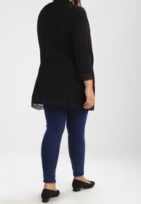 Maglietta nera a maniche lunghe con orlo in pizzo, abbinata a jeans skinny blu scuro e ballerine nere, mostrata da dietro.