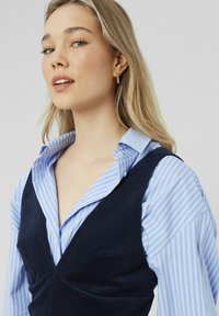 Un gilet en velours côtelé bleu marine superposé sur une chemise boutonnée rayée bleu clair. La chemise a un col classique et des manches retroussées. Boucles d'oreilles en métal doré.