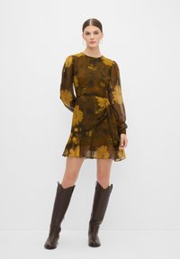 Korte bloemenjurk van bruine chiffon met gele bloemenpatronen, lange mouwen, een tailleband en een gerafelde zoom. Draagt met bruin kniehoge laarzen.
