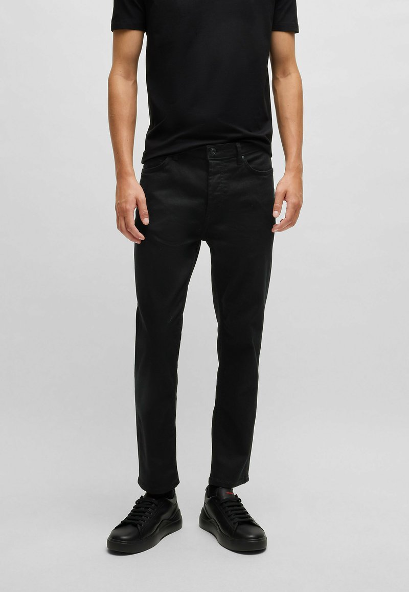 Sorte denimbukser med lige ben-design, der har diskret stikninger, fem lommer, og som er parret med sorte sneakers.