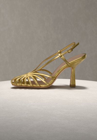 Χρυσό σανδάλι με λεπτά λουριά, ανοιχτό μπροστά με ψηλό τακούνι, διαθέτει λουρί πίσω με αγκράφα, εμφανίζεται πάνω σε στρογγυλή φωτεινή επιφάνεια με ουδέτερο φόντο.