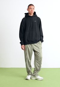 Ung mand står med hænderne i lommerne, iført sort Nike hættetrøje, lysegrå træningsbukser og hvide sneakers mod en ensfarvet hvid baggrund.