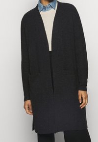 Dlouhý černý kardigan vyrobený z texturované pleteniny, s otevřeným předním dílem, dvěma předními kapsami a žebrovanými manžetami. Nosič je oblečený přes světlý svetr.